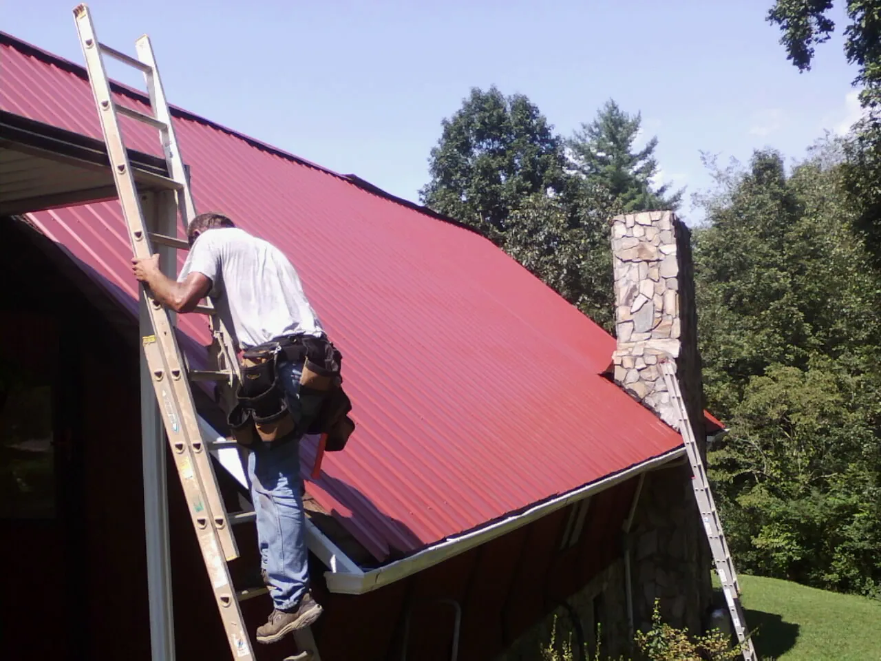 Roofer on a ladder beside a red metal roof and stone chimney — Soffit & Fascia Repair by Roofing Brothers in Wills Point